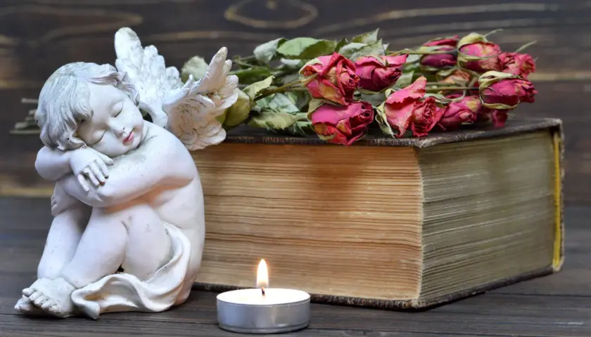 Figurka aniołka, stara księga, świeca i uschnięte róże na drewnianym tle.